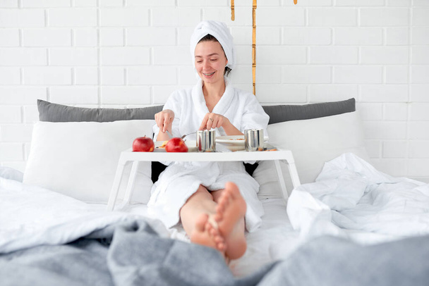 Happy girl has breakfast in bed in the bedroom with white walls - Φωτογραφία, εικόνα