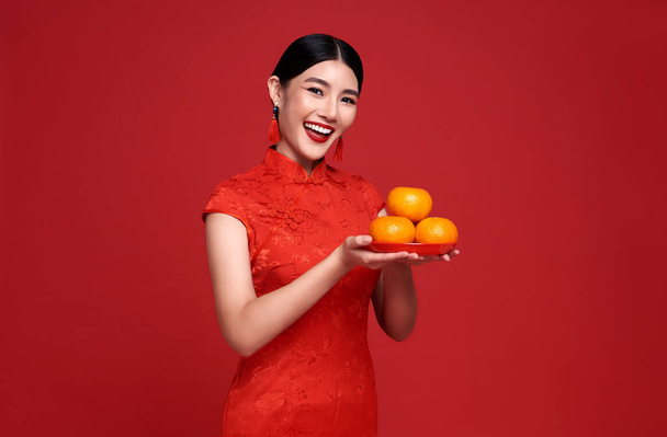 Happy Chinese new year. Asian woman wearing traditional cheongsam qipao dress holding fresh oranges isolated on red background. - Photo, Image