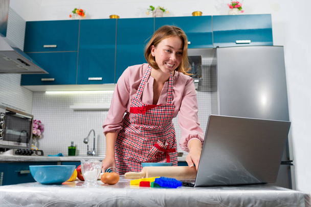Portrait of a young happy woman cooks in the kitchen using online recipes on her laptop. Indoors. Bottom view. The concept of cooking at home using online recipes and video communication. - Photo, Image