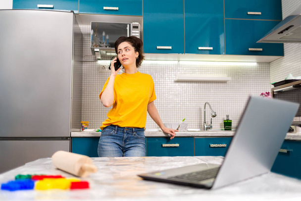 A young beautiful woman communicates on a smartphone in the home kitchen. In the foreground, a blurry view shows a laptop on a table. The concept of communication and of cooking at home. - Foto, Bild
