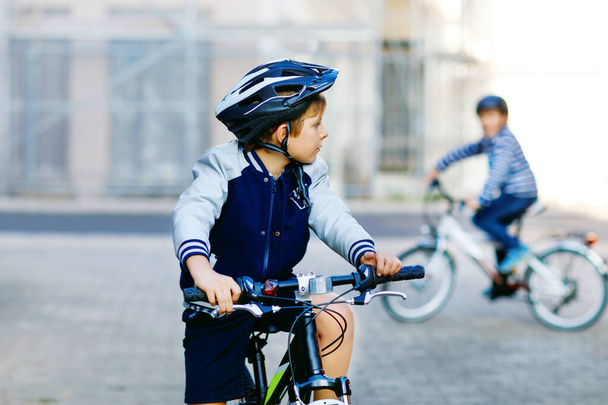 Двое школьников в защитном шлеме едут на велосипеде по городу с рюкзаками. Счастливые дети в красочной одежде катаются на велосипедах по дороге в школу. Безопасный способ для детей на открытом воздухе в школу - Фото, изображение