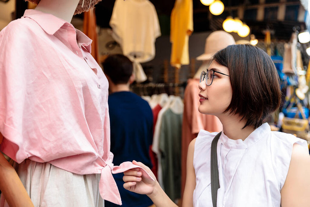 Azji młody krótkie włosy piękna kobieta nosi okulary patrząc różowy koszula na wyświetlaczu manekina na sprzedaż na rynku ulicy - Zdjęcie, obraz