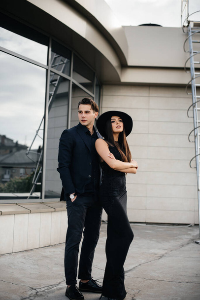 A beautiful, stylish pair of young people in black clothes and glasses stand against the background of an office building in the sunset. Fashion and style - Foto, afbeelding