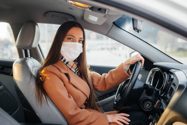A young girl sits behind the wheel in the car in the mask during the global pandemic and coronavirus. Quarantine - Photo, Image