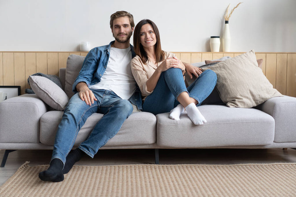 Happy young Caucasian couple relax on cozy sofa at home hugging look in distance dreaming together - Photo, Image