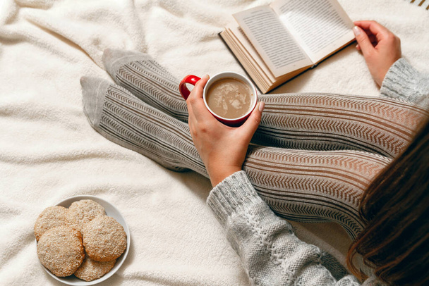 A young woman in a sweater and stockings, with biscuits and red coffee cup in hands relaxing in bed. top view - Foto, immagini