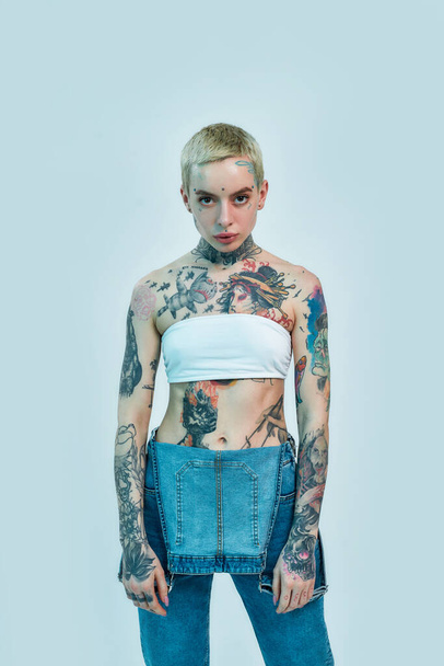Tattoo and piercing. A white woman with piercing and tattoos standing in front of a camera looking into it wearing a denim overall - Fotografie, Obrázek