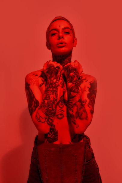 Tattoo and piercing. A white woman with tattoos and piercing topless wearing denim overall with her hands covering breasts standing looking into a camera - Φωτογραφία, εικόνα