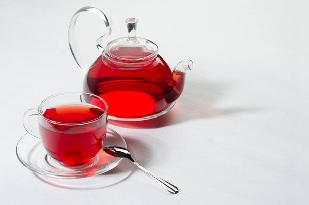 Red hibiscus tea in transparent teapot with cup in sunlight with shadow on white wood board, copy space. - Foto, Imagem