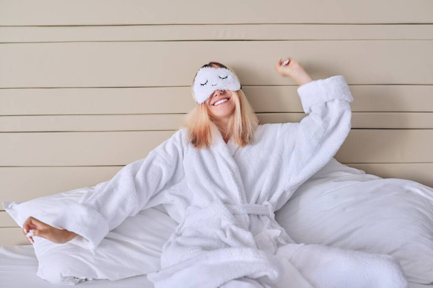 Young woman in white bed in bathrobe, eye mask, female yawns - Fotografie, Obrázek