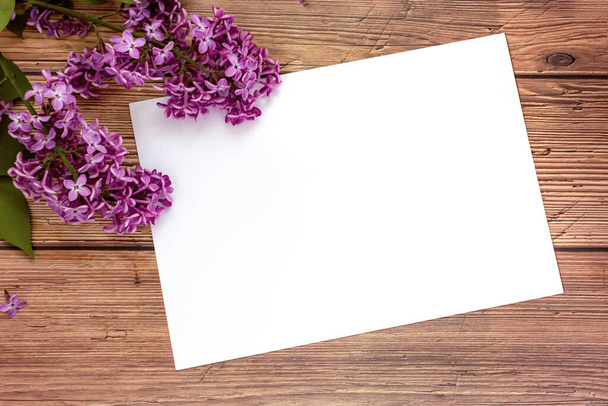 White blank sheet of paper with blooming lilac flowers on wooden background. Invitation or greeting card for Valentine's Day and Mother's Day. Top view, flat lay, mock up, copy space. Spring concept. - Foto, imagen