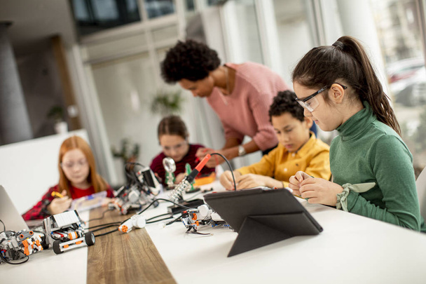 Groep gelukkige kinderen met hun Afro-Amerikaanse vrouwelijke wetenschap leraar met laptop programmeren elektrisch speelgoed en robots op robotica klaslokaal - Foto, afbeelding