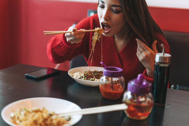 Krásná usmívající se mladá asijská žena v červeném oblečení jíst nudle s bambusovými hůlkami v čínské vietnamské restauraci - Fotografie, Obrázek