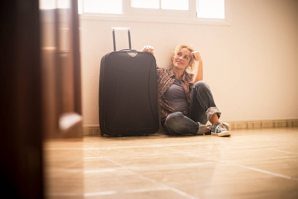 Λευκή γυναίκα ξάπλωσε στο πάτωμα ενός άδειου δωματίου χαμογελώντας και χαλαρώνοντας με μεγάλες αποσκευές - Φωτογραφία, εικόνα