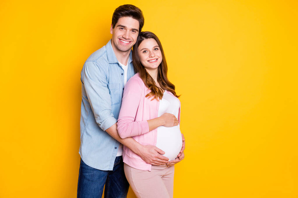 Photo of optimistic cute young couple stand hug pregnant woman wear casual outfits isolated on yellow color background - Zdjęcie, obraz