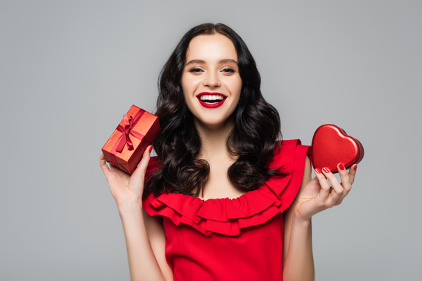 cheerful woman with red lips holding heart-shaped gift box and wrapped present with bow isolated on grey - Photo, Image