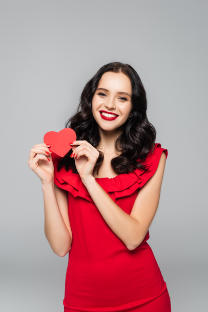 cheerful young woman in dress holding red paper heart isolated on grey - 写真・画像
