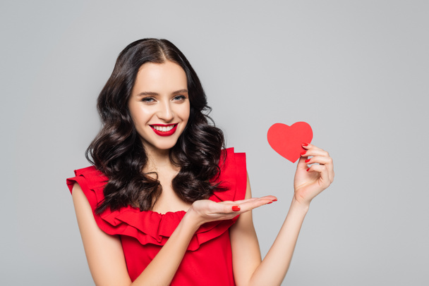 smiling young woman pointing with hand at red paper heart isolated on grey - Photo, Image