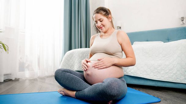 Portrait de belle femme enceinte caressant son gros ventre après avoir pratiqué le yoga et la forme physique sur tapis à la maison. Concept de mode de vie sain, de santé et de sport pendant la grossesse - Photo, image
