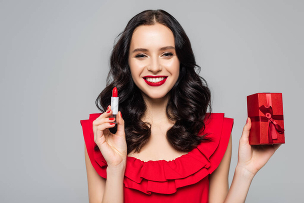 cheerful young woman holding red lipstick and wrapped present isolated on grey - Фото, изображение