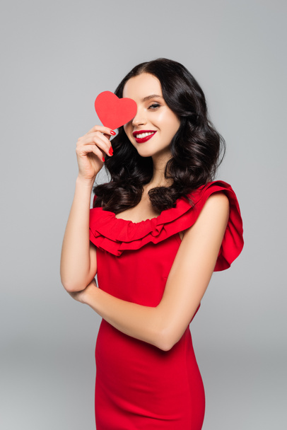 smiling young woman covering eye with red paper heart isolated on grey - Foto, Imagem