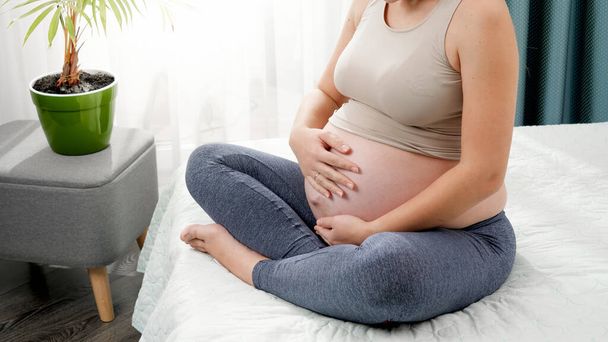Beautiful young pregnant woman in leggings sitting on bed side and stroking her big belly against bright morning sun shining through window. Concept of healthy lifestyle, healthcare and sports during - Photo, image
