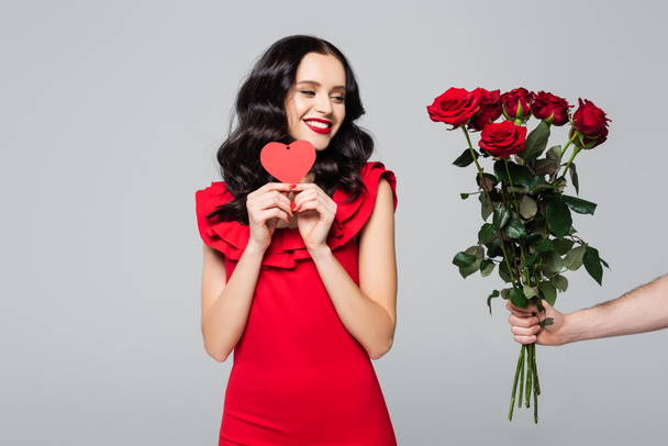 άντρας που δίνει τριαντάφυλλα σε ευτυχισμένη γυναίκα με κόκκινο φόρεμα κρατώντας μια κάρτα σε σχήμα καρδιάς που απομονώνεται στο γκρι - Φωτογραφία, εικόνα