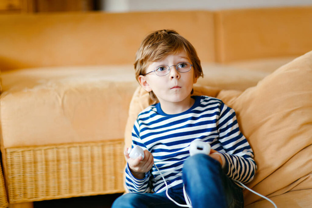 Cute little blond kid boy with glasses playing with a video game console. Child having fun at home during corona virus lockdown quarantine. Lonely alone boy without friends, indoors - 写真・画像