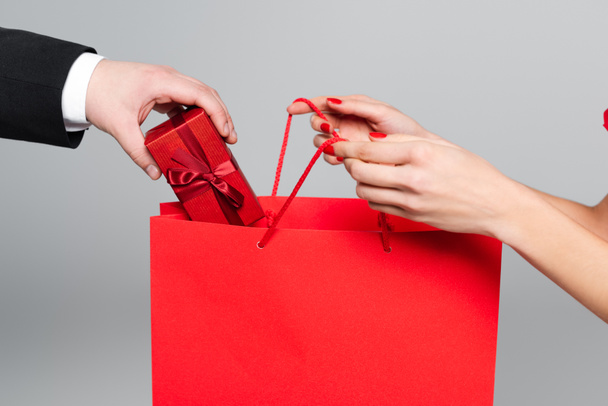 cropped view of man putting gift box in paper bag in hands of woman with red lips isolated on grey - Photo, Image