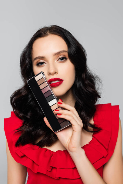 young brunette woman holding eye shadow palette isolated on grey - Fotografie, Obrázek