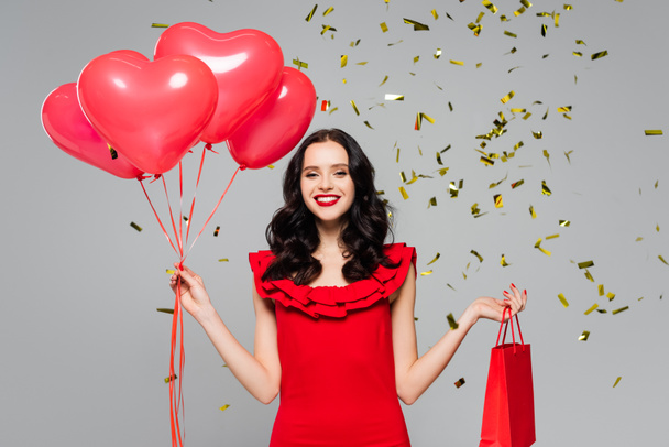happy woman holding red heart-shaped balloons and shopping bag near falling confetti on grey - Foto, imagen