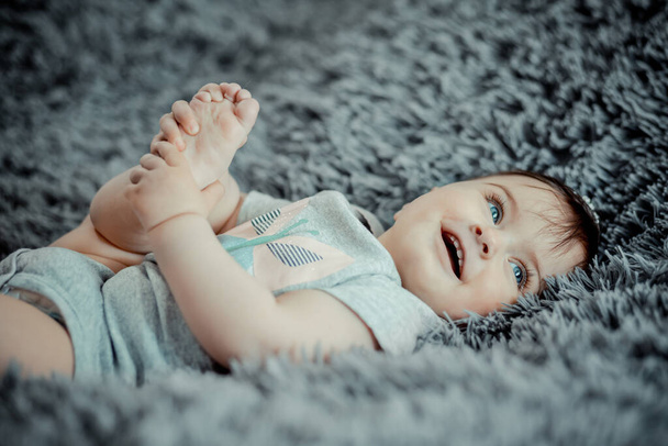 Heureux gai bébé de sept mois tenant sa jambe et couché sur une feuille molle grise - Photo, image