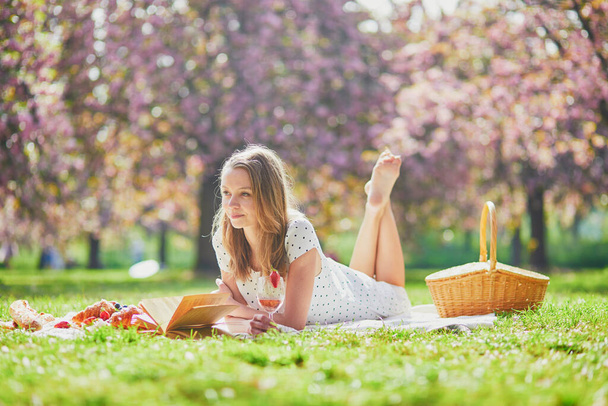 Beautiful young woman having picnic on sunny spring day in park during cherry blossom season - Foto, afbeelding