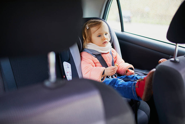 Adorable toddler girl in modern car seat eating cookie. Little kid traveling by car. Child safety on the road. Trip with a baby - Photo, Image