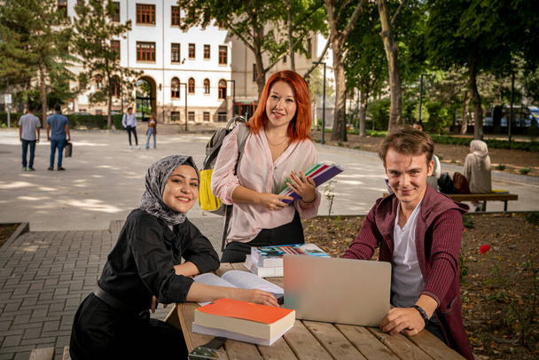 Νέοι μαθητές μελετούν μαζί, συνομιλούν, κοιτάζουν τον υπολογιστή, βλέπουν ταινίες και γελούν στον κήπο του σχολείου. Πανεπιστήμιο, πανεπιστημιούπολη, σχολικός κήπος, μουσουλμάνος φοιτητής, χριστιανός, εβραίος, διαφορετικοί άνθρωποι - Φωτογραφία, εικόνα