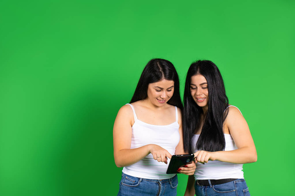 The twin sisters smile and stare at the tablet against a green background with an empty side space. - Foto, Imagen