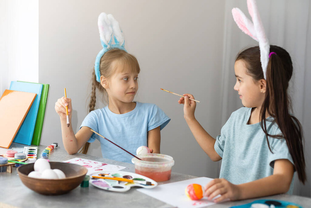 Veselé Velikonoce. Dvě sestry malují velikonoční vajíčka. Šťastné rodinné děti se připravují na Velikonoce. Roztomilé malé dítě dívka nosí králičí uši na velikonoční den. - Fotografie, Obrázek