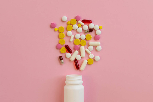Pilules médicales assorties et capsules dispersées à partir d'une bouteille en plastique blanc sur un fond rose. Concept de traitement pour une personne malade. Vue de dessus. Pose plate. - Photo, image