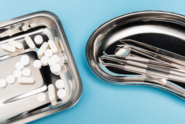 Hochwinkelansicht von Pillen und zahnärztlichen Werkzeugen auf Tabletts auf blauem Hintergrund - Foto, Bild