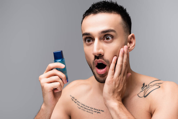 shocked hispanic man touching face while holding bottle of aftershave lotion isolated on grey - Foto, Imagem