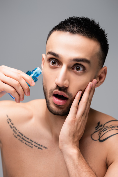 asombrado hombre hispano tocando la cara mientras se aplica loción aftershave aislado en gris - Foto, imagen