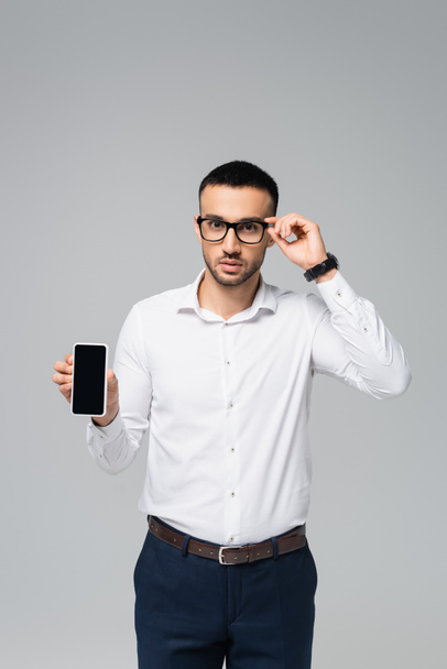 gerente hispânico ajustando óculos enquanto mostra celular com tela em branco isolado em cinza - Foto, Imagem