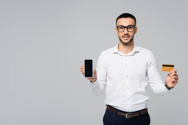 cheerful hispanic businessman holding credit card and smartphone with blank screen isolated on grey - 写真・画像