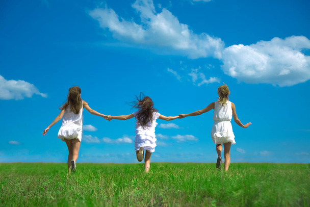  running girls  at green grass at background of blue sky with clouds.  young adult women  on summer field. Empty space for inscription. Back view.  - Foto, Bild