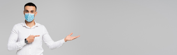 Ισπανόφωνος επιχειρηματίας με λευκό πουκάμισο να δείχνει στην άκρη, ενώ κοιτάζοντας κάμερα απομονωμένη σε γκρι, banner - Φωτογραφία, εικόνα