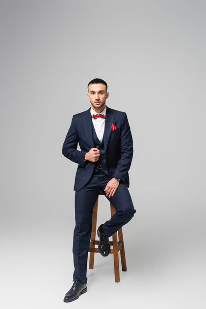 full length view of young hispanic man in blue suit posing near high stool on grey - Foto, imagen