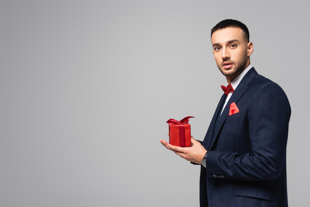 brunette hispanic man in elegant suit holding valentines day present and looking at camera isolated on grey - Foto, afbeelding