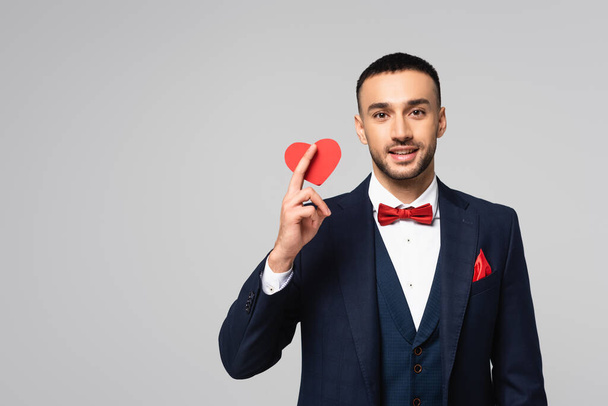 heureux hispanique homme en costume élégant tenant rouge coeur coupé en papier isolé sur gris - Photo, image