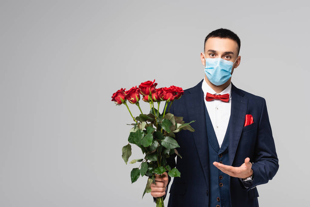 młody Hiszpan w niebieskim garniturze i maska medyczna wskazująca na czerwone róże odizolowane na szaro - Zdjęcie, obraz