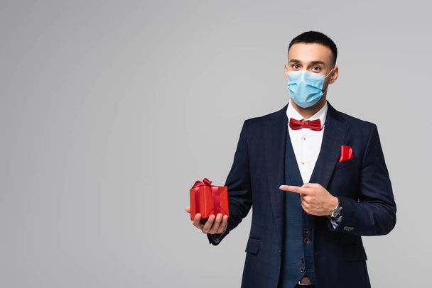 young hispanic man in elegant suit and medical mask pointing at red gift box isolated on grey - Foto, Bild